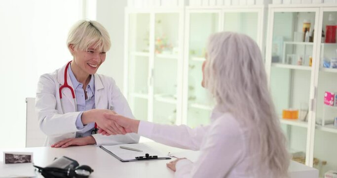 Smiling female doctor shakes hand of mature patient sitting at table. Women joyfully greet each other at consultation with specialist slow motion