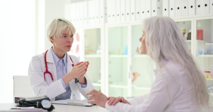 Doctor clasping hands together talks about treatment of senior lady patient. Mature woman listens attentively to specialist sitting at table slow motion