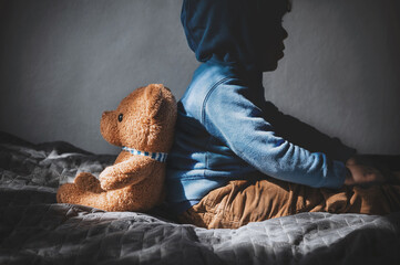 A sad little boy sits back to back with a teddy bear. Lonely depressed child in a room with a teddy bear. Post-traumatic disorder in a child