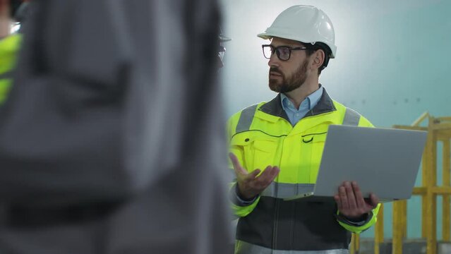 Industrial factory female and male engineers holding laptop. Bearded worker in hard hat and yellow vest has discussion with project manager. Two employees work and communicate at construction site.