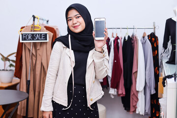 Young Asian muslim woman holding phone with blank screen at her clothing shop