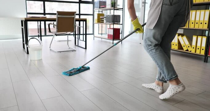 Maid does industrial cleaning in modern office at end of working day. Woman in rubber gloves washes wooden floor with mop and detergent slow motion