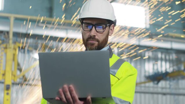 Thoughtfull mature engineer is holding laptop. Male looking at work process. Worker at backgrounf of welding sparks flying. Construction builder dressed up in reflective vest and casque.