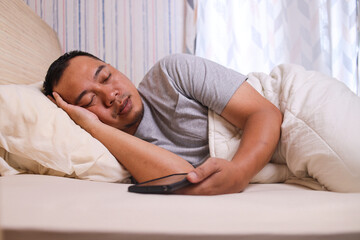 Young Asian man sleeping spread out in bed with his smartphone. Sleep position concept.