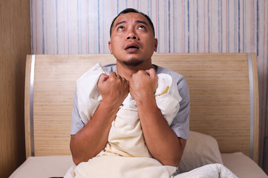 Asian man sitting in bed stressed and scared suffering from nightmare and horror bad dream, grabbing pillow frightened and paranoid in sleeping disorder and resting problem