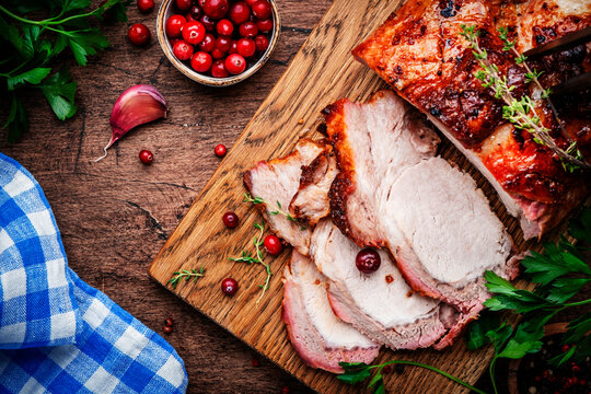Baked pork loin on rustic wooden cutting board with spices, herbs and cranberries. Wood kitchen table background, top view