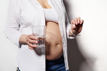 Close-up pregnant woman holding glass of water and medical pill with vitamins and minerals to take during pregnancy, posing bare belly, isolated white background. Healthcare and medicine. Gynecology