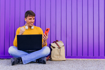 Smiling male student using smart phone over purple wall of university campus.
