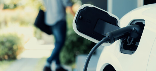 Focus electric car charging at home charging station with blurred progressive man walking in the...