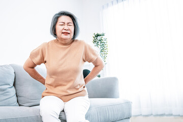 An agonizing senior woman experiencing back pain while sitting on her sofa at home and holding her...