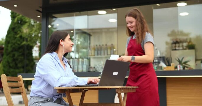 Young attractive business woman with coffe in trendy suit using wireless laptop for work outdoors.At one point the barista comes and serves her coffee.