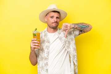 Young caucasian man holding a cocktail isolated on yellow background showing thumb down with negative expression