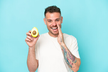 Young caucasian man holding an avocado isolated on blue background shouting with mouth wide open