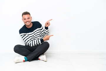 Young man sitting on the floor isolated on white background pointing finger to the side and presenting a product