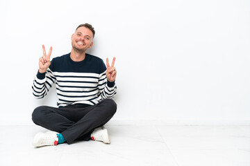 Young man sitting on the floor isolated on white background showing victory sign with both hands