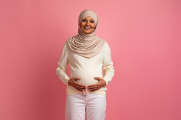 Beautiful Muslim Pregnant Woman Tenderly Embracing Belly While Posing Against Pink Background