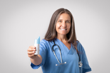 Happy senior woman doctor in uniform recommending at camera nasal wash bottle for spray medicine...