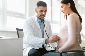 Happy pregnant woman visiting doctor, male practitioner examining pregnant smiling lady, listening to baby's heartbeat