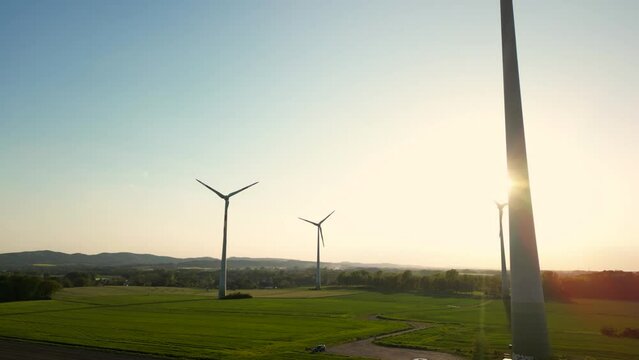 Large wind turbines with blades in field aerial view bright orange sunset blue sky wind park slow motion drone turn. Silhouettes windmills, large orange sun disc summer lens flare. Alternative energy