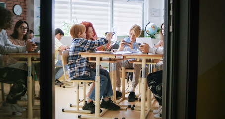 View from door of classroom on Caucasian kids and teenagers sitting at desk and studying together. Classes for kids after lessons. Indoors. Children of diferent ages cooperating and coworking.