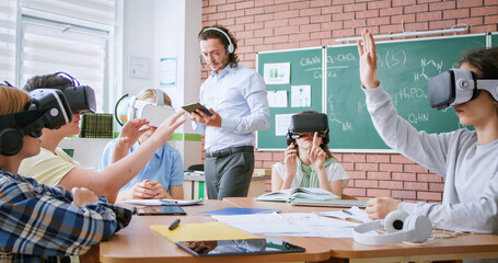 Group of young pupils in VR headsets sitting together at desk while male teacher with digital tablet in hands explaining education program. Modern technologies during interactive lesson at school.