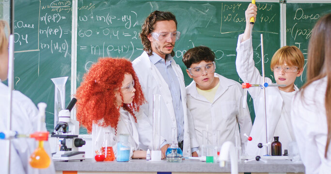 Children analyzing and mixing liquid in test tube with help of male teacher at school. Caucasian man and pupils standing together in classroom and wearing protective glasses with white lab coats.