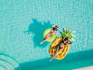 Top view of beautiful girls in Santa's hats relaxing on inflatable pineapple mattress in pool. Celebrating Happy New year on resort.