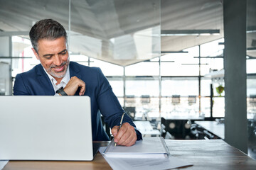 Busy happy middle aged smiling professional business man company executive ceo manager or lawyer wearing suit sitting at desk in modern office working on laptop computer writing notes, copy space.