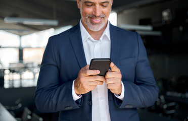 Happy mid aged business man ceo wearing blue suit standing in office using cell phone. Mature businessman professional manager holding mobile checking corporate apps on cellphone. Close up