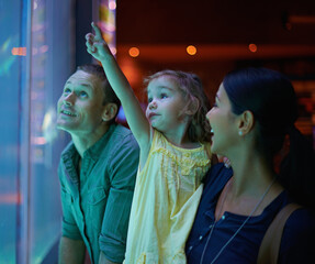 Family, aquarium and child pointing at fish for learning, curiosity or knowledge, bonding or care. Mother, fishtank and happy kid with father watching marine animals swim underwater in oceanarium.