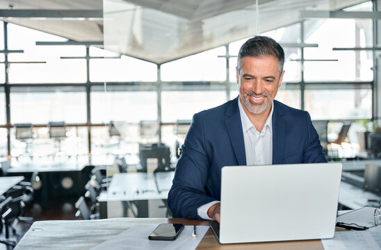 Smiling mature adult business man executive sitting at desk using laptop. Happy busy professional mid aged businessman ceo manager working on computer corporate technology in modern office.