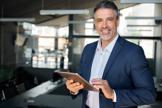 Smiling mature professional business man, happy middle aged successful company ceo, confident executive wearing blue suit standing in office holding tablet computer corporate technology. Portrait