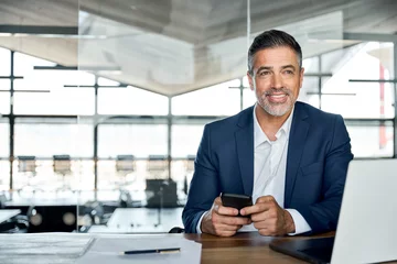 Gardinen Smiling mid aged business man ceo wearing blue suit sitting in office using cell phone solutions. Mature businessman professional executive holding mobile working at desk with laptop and smartphone. © insta_photos