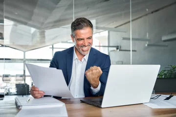 Fototapeten Happy mature older business man ceo wearing suit celebrating success at work in office holding papers looking at laptop rejoicing company growth, goals achievement good results screaming yes. © insta_photos