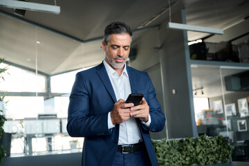 Fototapeta na wymiar Confident mature business man ceo wearing blue suit standing in office using cell phone. Mid aged businessman professional executive holding mobile smartphone working, texting messages on tech device.