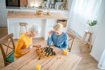 two women mature senior females sisters play chess board game at home