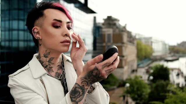 An non-binary androgynous person is doing make-up outdoors to himself.