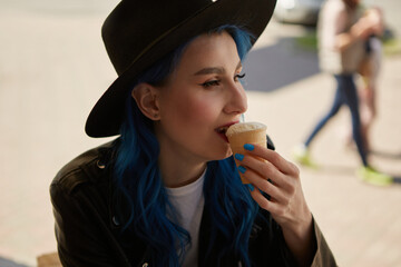 Portrait of a beautiful diverse woman with dyed blue hair eating an ice cream outdoor. Attractive white female person wearing stylish hipster hat and leather jacket enjoying the dessert in a cafe