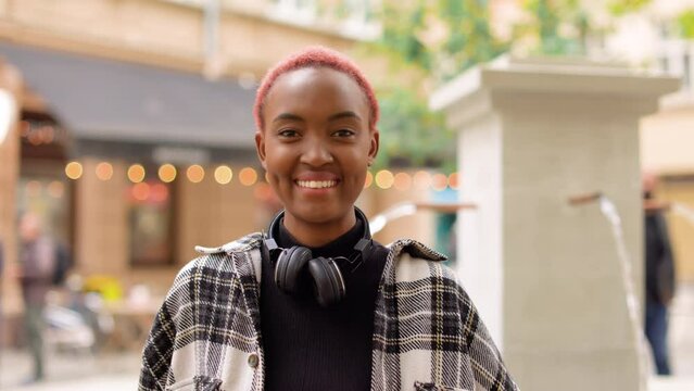 Close up of young cheerful African American woman with headphones standing near small fountain and smiling at camera outdoor. Positive emotions. Beautiful stylish female with smile on face