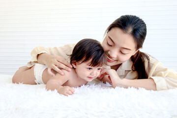Portrait of happy toddle baby lying on fluffy white rug with mother, little cute kid girl tries to crawl, mom hugs her daughter child, love in family, childhood and motherhood concept.