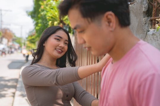 An asian lady wearing a brown long sleeves top is trying to console the guy in pick shirt by putting her hand in his shoulder and smiling.