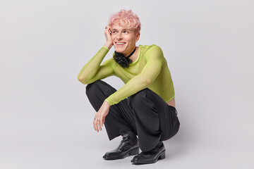 Cheerful dreamy pink haired transgender man squats indoor against white wall being in good mood...