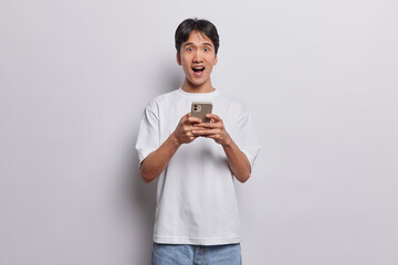 Horizontal shot of shocked young Japanese man surprised by mobile application expresses amazement stands with widely opened mouth dressed in t shirt and jeans isolated over white background.