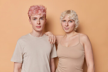 Portrait of girlfriend and boyfriend stand closely to each other dressed in casual t shirts look directly at camera have good relationship pose for making photo isolated over brown background