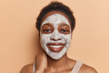 Portrait of cheerful African woman applies white nourishing mask on face to refine pores provides even skin tone touches neck smiles broadly looks directly at camera isolated over brown background
