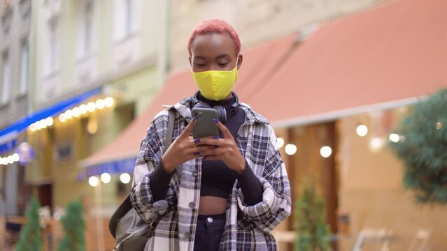 Portrait of beautiful African American female tourist standing in city on street with cafeterias wearing face mask texting on smartphone. Young stylish female using social media on cellphone outdoor