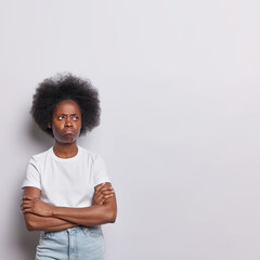 Vertical shot of displeased African woman with curly hair stands in thoughtful expression keeps arms folded concentrated aside has discontent expression dressed in t shirt and jeans white background