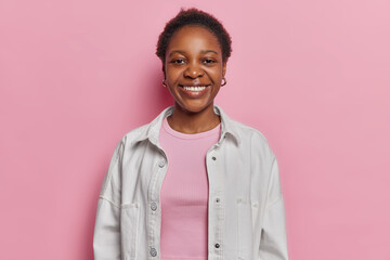 Studio shot of dark skinned woman smiles broadly has perfect white teeth glad to hear something interesting dressed in white jacket isolated over pink background. People and positive emotions concept