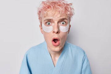 Shocked pink haired man applies beauty patches under eyes for reducing wrinkles keeps mouth opened dressed in blue robe undergoes skin care procedues has speechless expression isolated over white wall