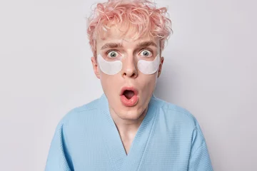Zelfklevend Fotobehang Shocked pink haired man applies beauty patches under eyes for reducing wrinkles keeps mouth opened dressed in blue robe undergoes skin care procedues has speechless expression isolated over white wall © Wayhome Studio